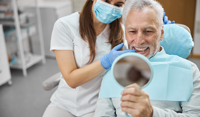 Older man sitting in a dental chair looking in a handheld mirror at his new dental implants from Aesthetic Periodontal & Implant Specialists in San Antonio, TX